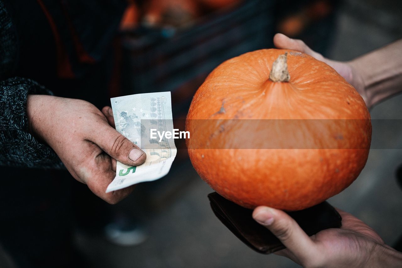Cropped image of people exchanging pumpkin with paper currency outdoors