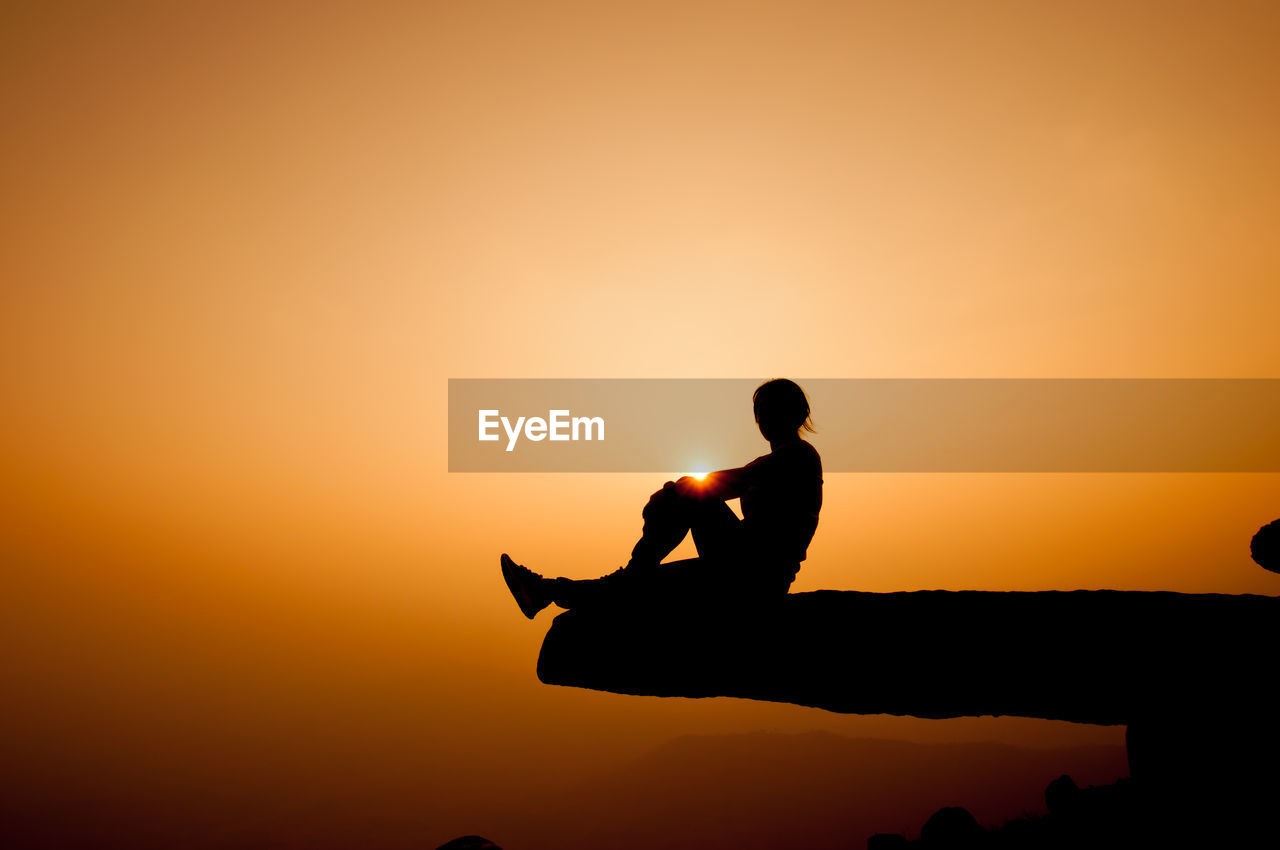 Silhouette of woman sitting on cliff against sky during sunset