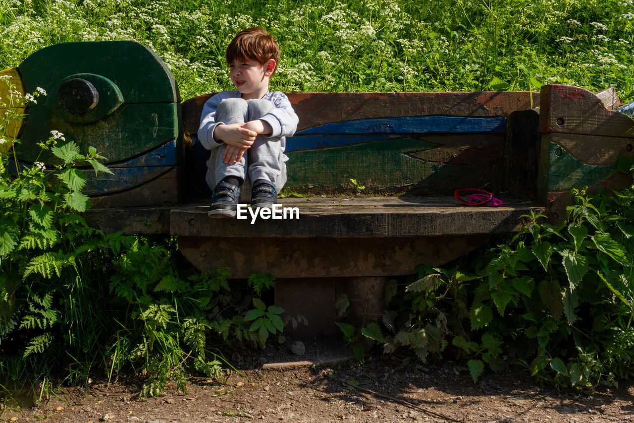 A cute redhead boy sitting on a bench in a park in front of green lush vegetation