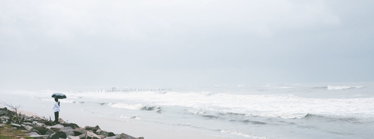 Panoramic shot of man with umbrella standing on rocks at seashore