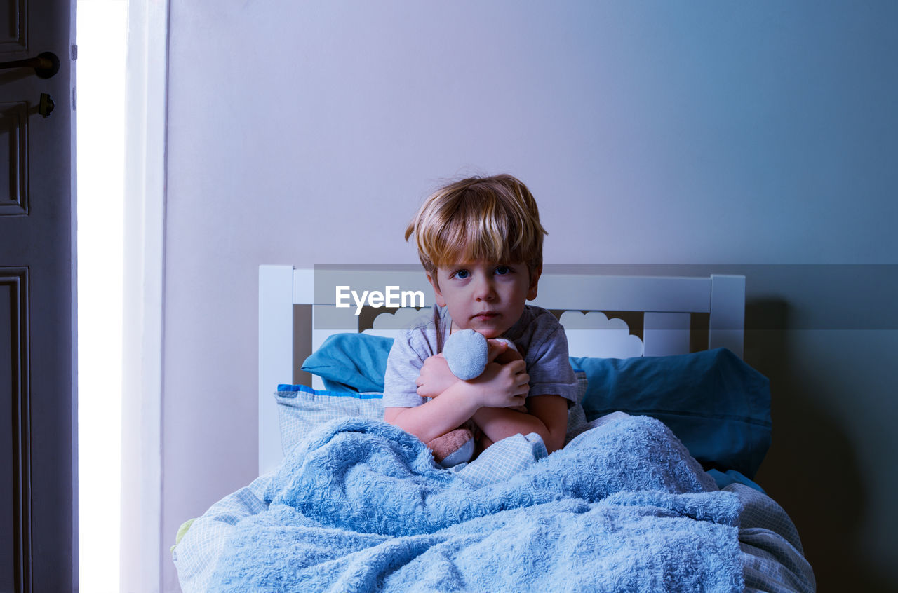 portrait of cute girl playing with stuffed toy while sitting on bed at home