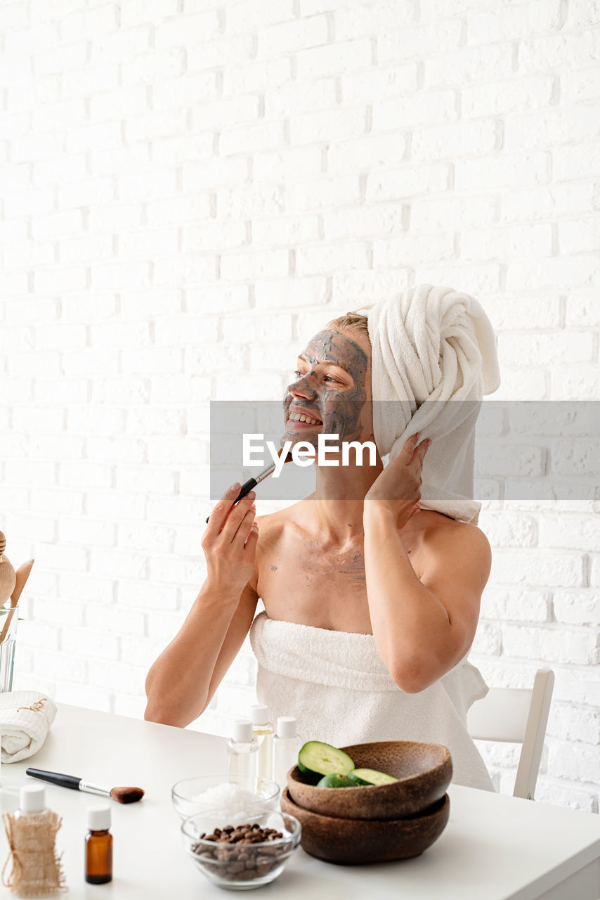 Young woman wearing white towels applying spa facial mask on her face with a cosmetic brush