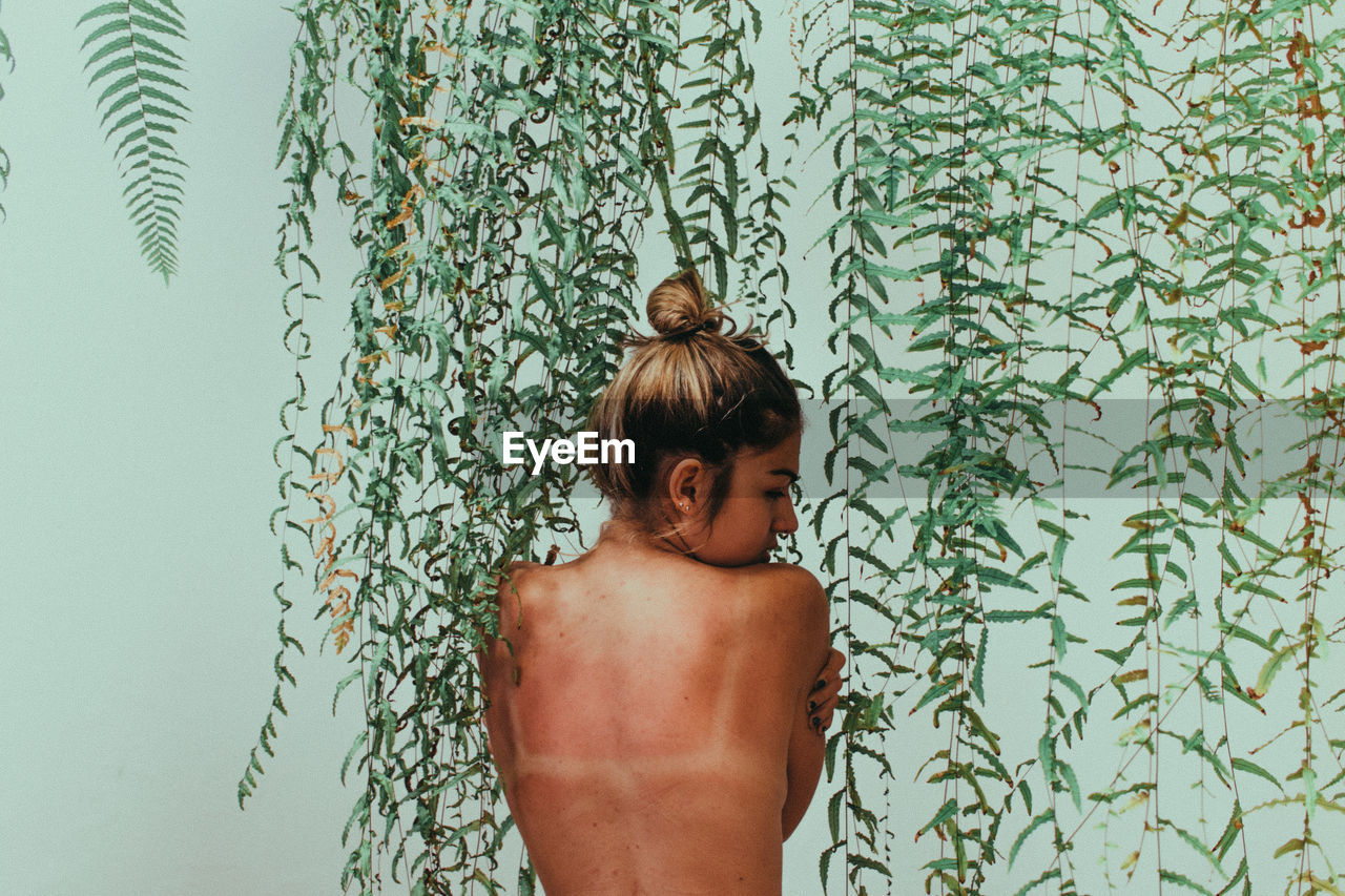 Rear view of shirtless woman standing against ivy wall