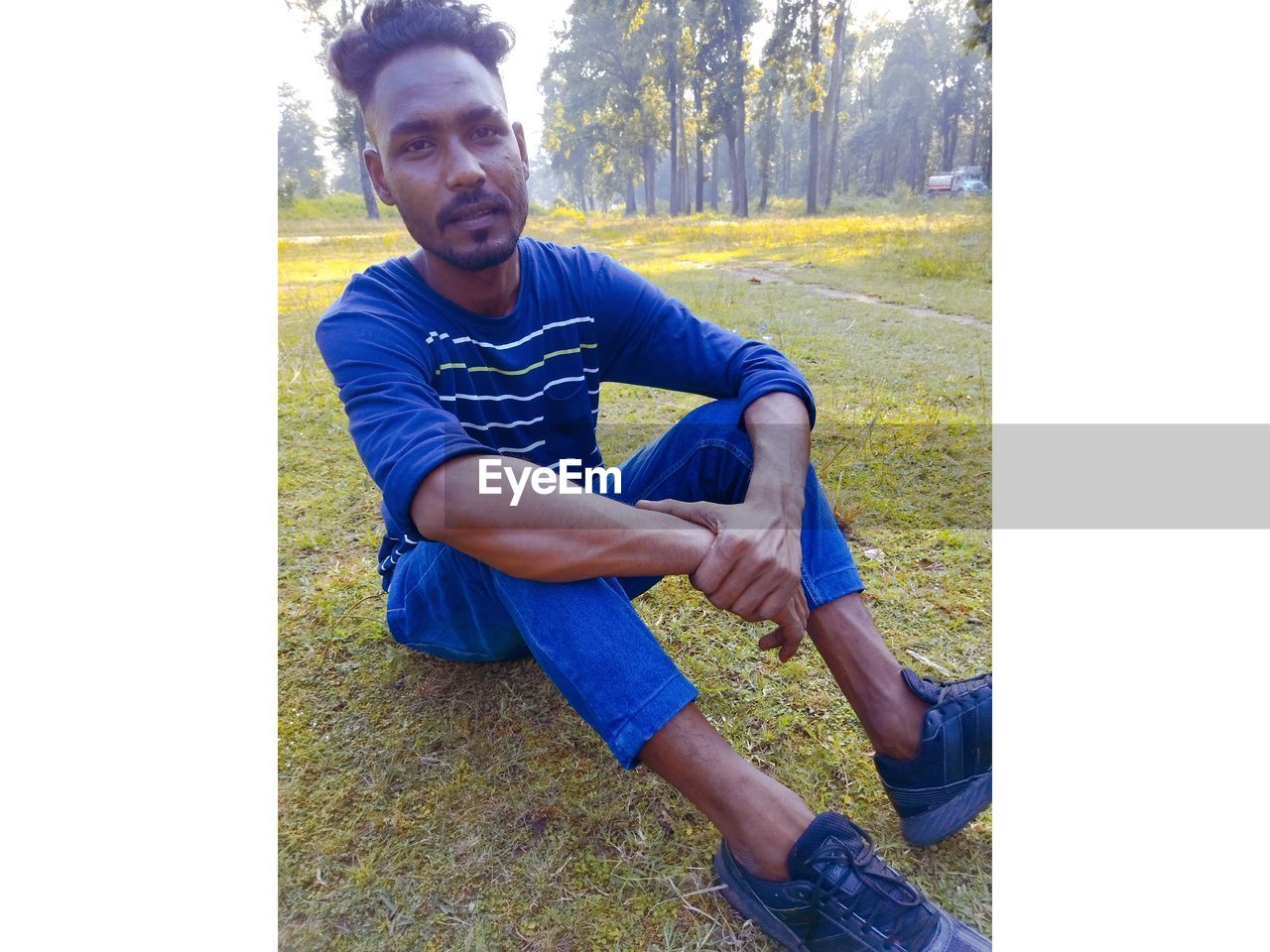 one person, casual clothing, sitting, full length, men, leisure activity, footwear, lifestyles, young adult, plant, adult, nature, day, grass, front view, looking at camera, portrait, person, tree, transfer print, relaxation, clothing, arm, auto post production filter, outdoors, beard, sports, hand, blue, human face, facial hair, smiling, electric blue, looking