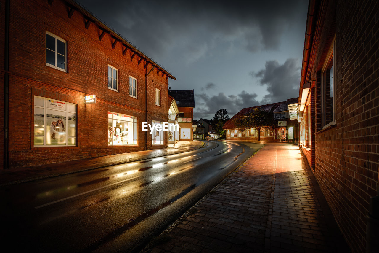 Wet street amidst illuminated buildings at night