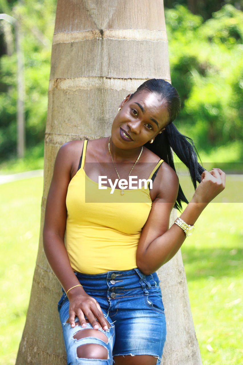 Portrait of smiling young woman standing by tree at park