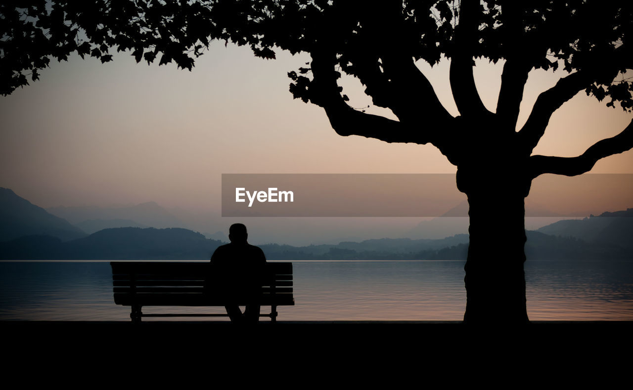 Silhouette man sitting on bench against lake at sunset