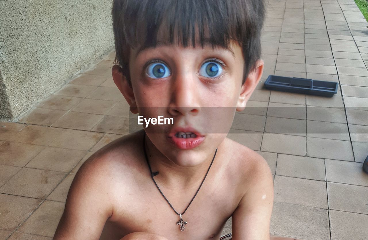 Close-up portrait of shirtless boy with raised eyebrows on tiled floor