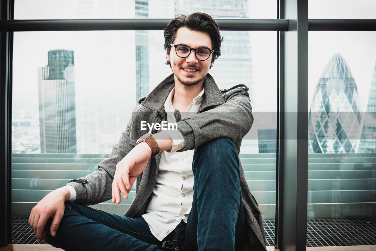 PORTRAIT OF SMILING YOUNG MAN SITTING BY WINDOW