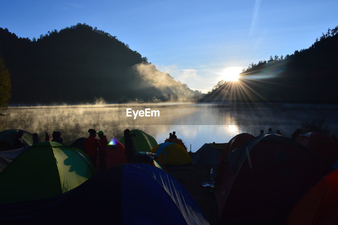 Group of people camping by lake in forest during sunset
