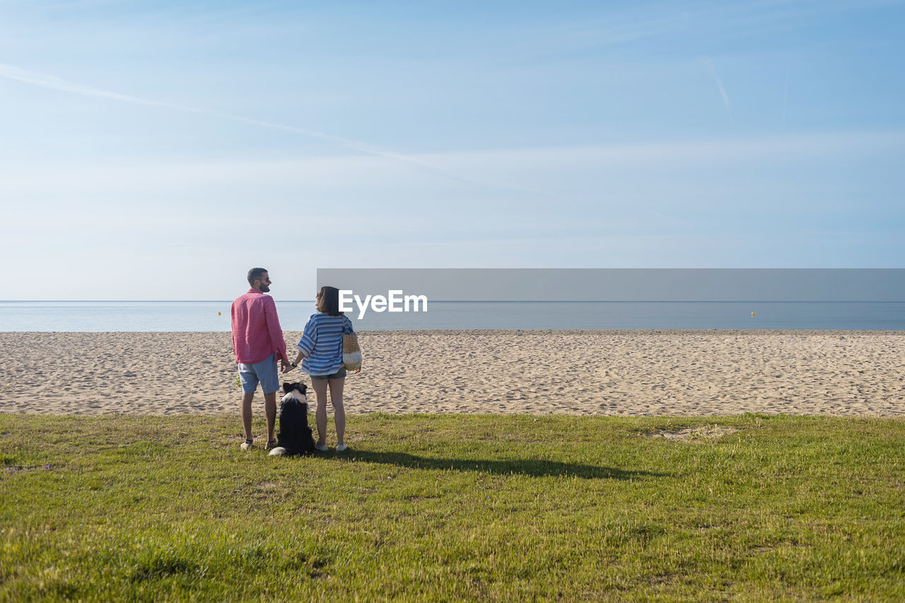 Back view of couple standing and holding hands near their dog on beach