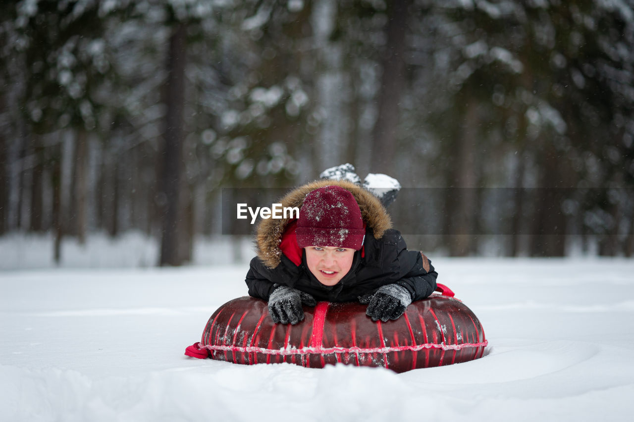 A smiling teenage boy in a black jacket with a fur and red hat lies on a tubing in a snow-covered