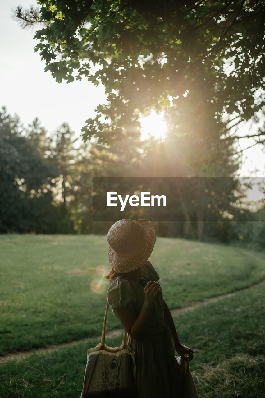 Rear view of woman standing on field against trees during sunset