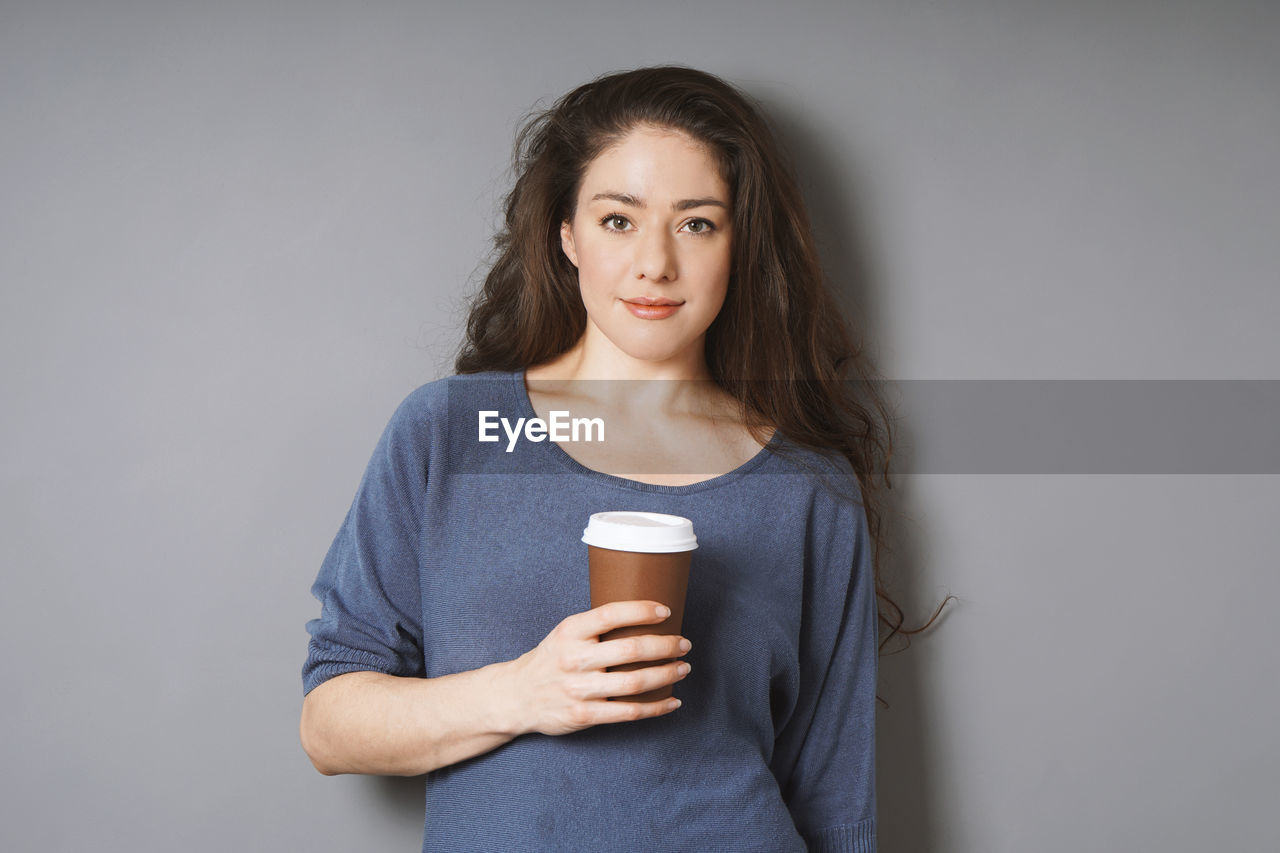 Portrait of young woman having coffee while standing against gray background