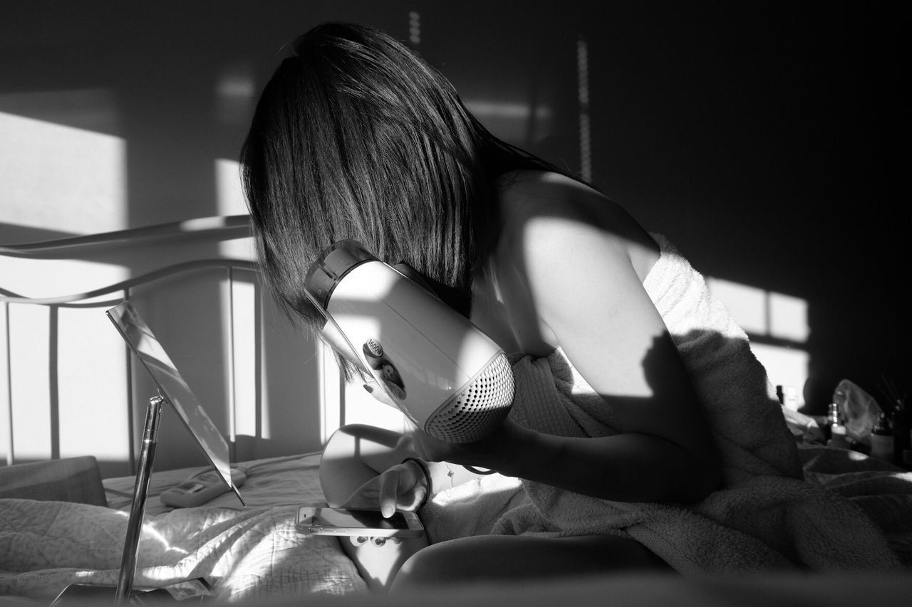 Woman wrapped in towel holding hair dryer while using phone on bed at home