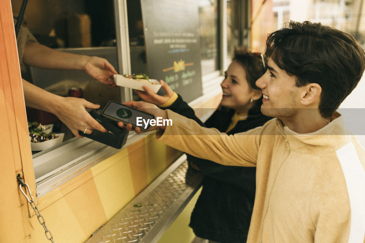 Smiling young man doing online payment while buying food from concession stand