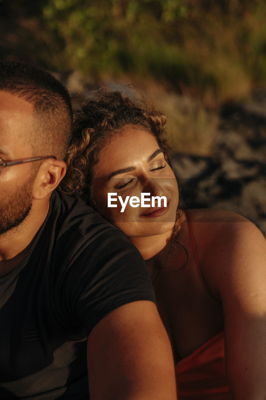 Smiling woman with eyes closed resting on boyfriend's shoulder during sunset