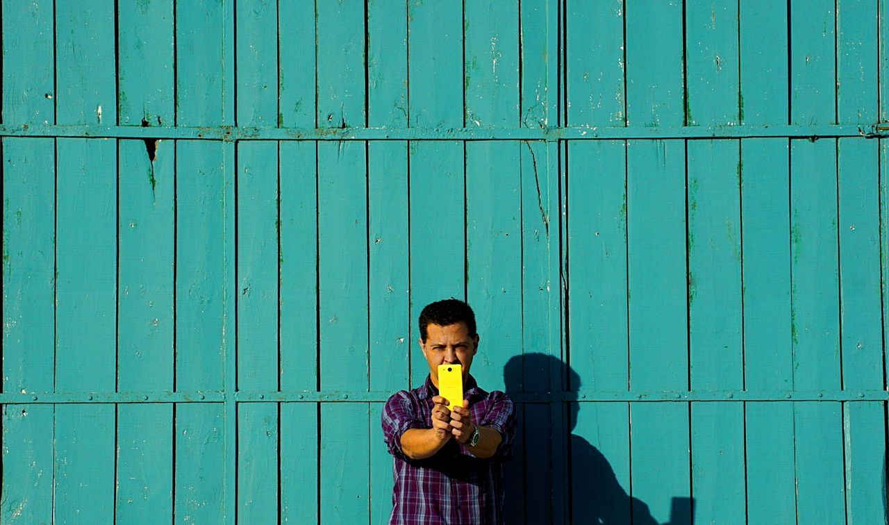 Man photographing through mobile phone while standing against blue wooden wall