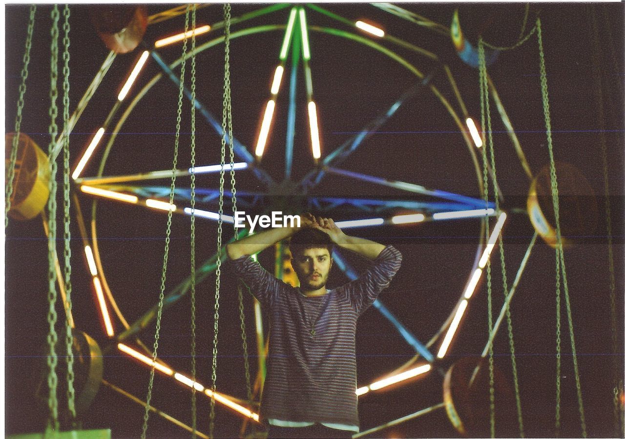 Portrait of a young man standing against illuminated ride