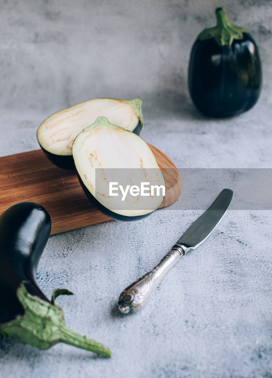 Halved eggplant or aubergines on chopping wooden board and knife over gray background.