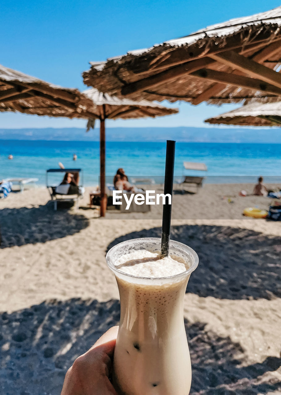 Personal perspective of person holding glass of cold iced coffee on beach in summer.