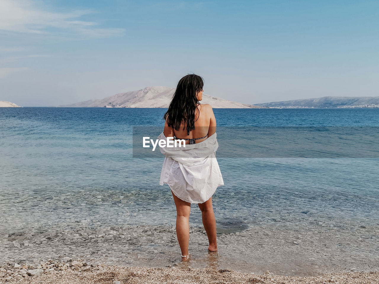 Rear view lifestyle image of stylish young woman in white shirt standing on beach and looking at sea