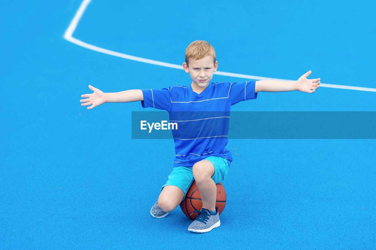 Portrait of boy with arms outstretched sitting on basketball at court