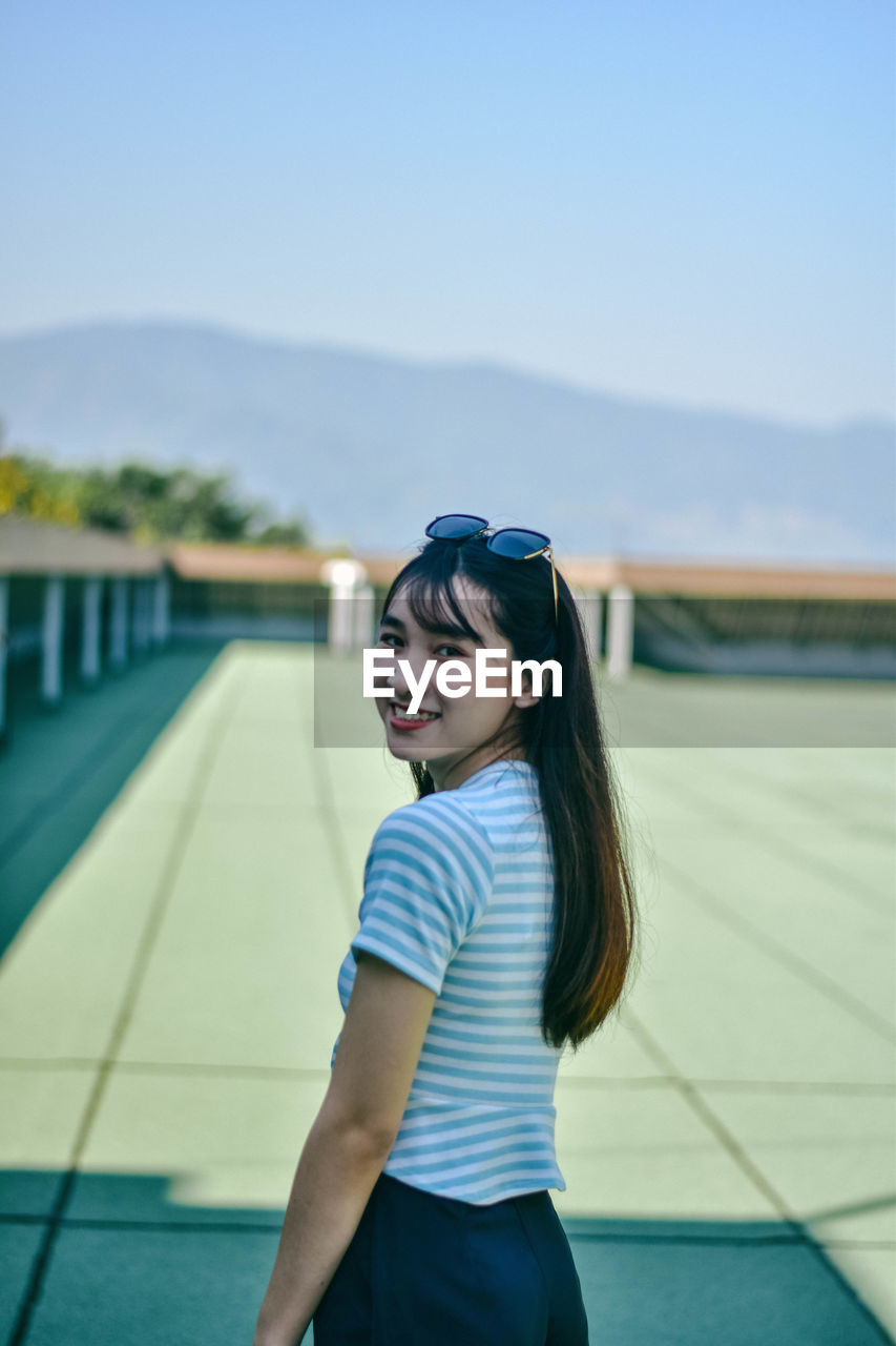 Portrait of cheerful young woman standing on building terrace against sky