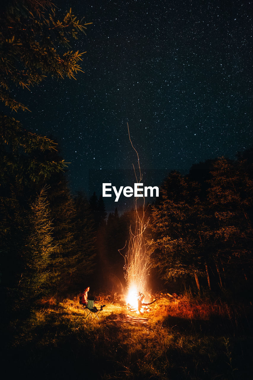 Woman sitting by fire on land against sky at night