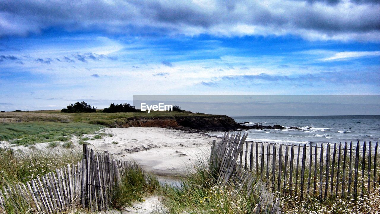 Wooden fence at beach against cloudy sky