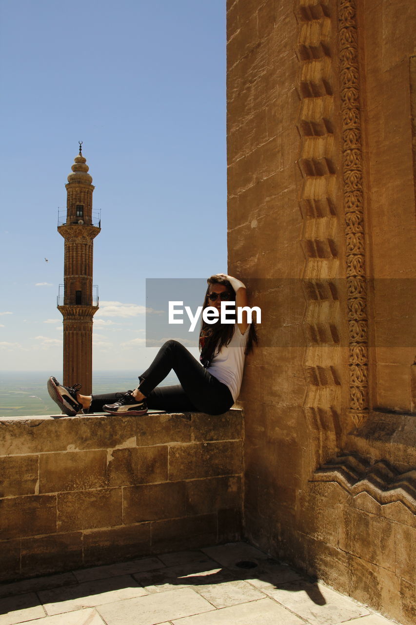 YOUNG WOMAN SITTING ON WALL AGAINST SKY WITH COLUMNS