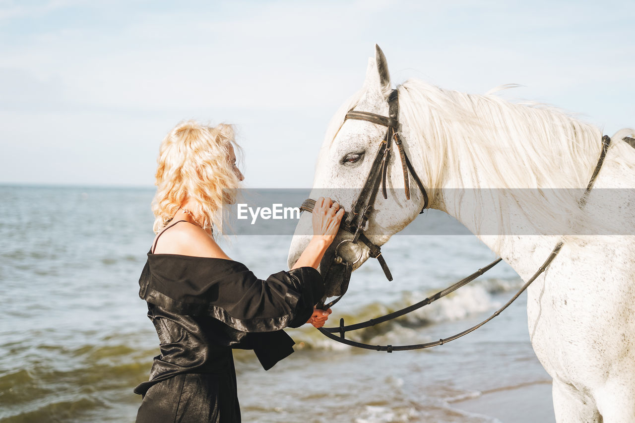 Young blonde woman in black clothes riding white horse on seascape background