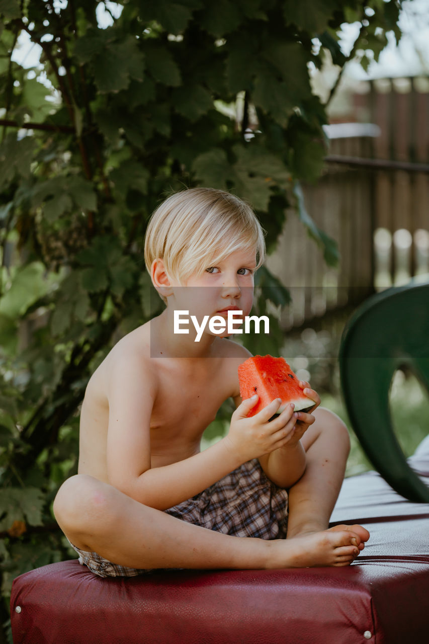 Portrait of shirtless boy holding food sitting outdoors