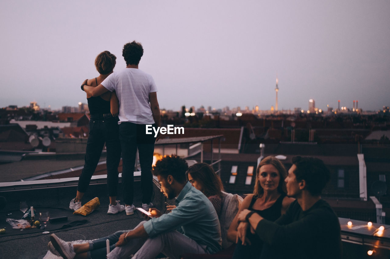 Couple looking at city while friends relaxing on terrace during rooftop party