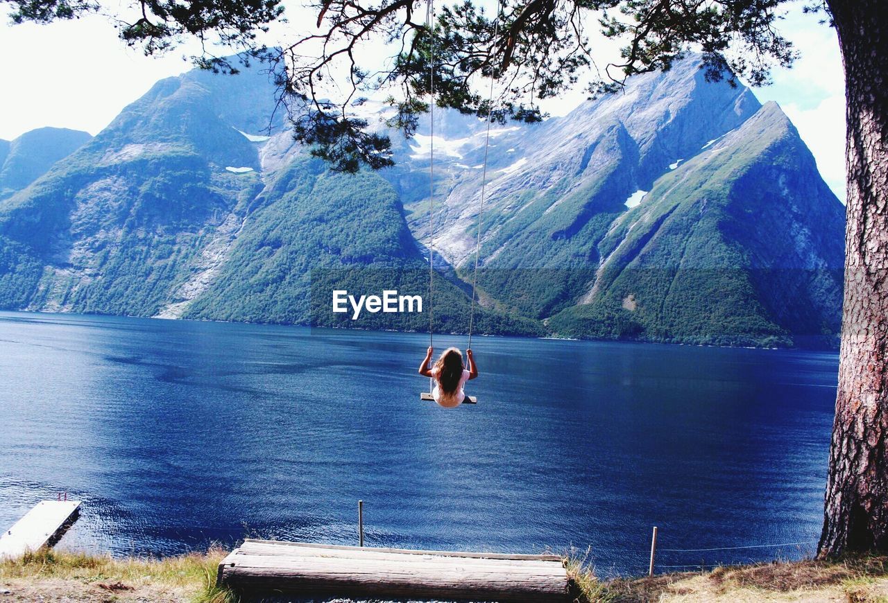 Rear view of girl swinging over sea against mountains