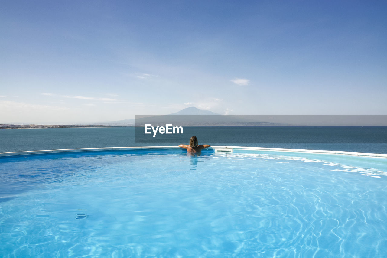 Adult woman relaxing in infinity pool looking at horizon with mount etna