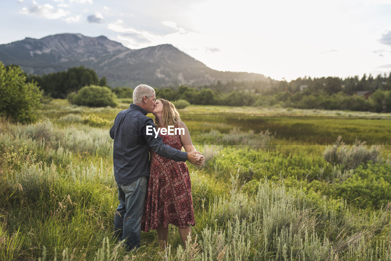 Romantic couple holding hands while kissing on grassy field against sky in forest