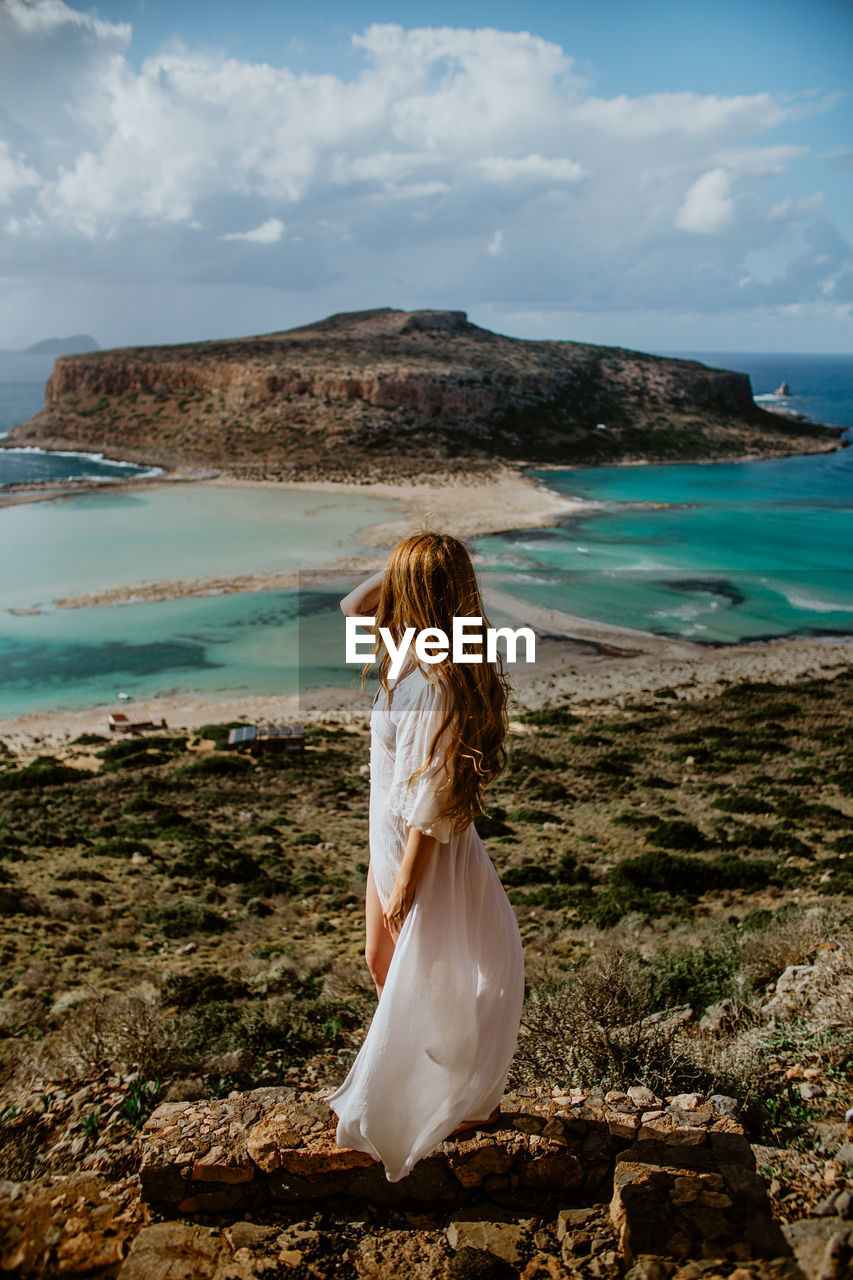 Side view anonymous female in white maxi beachwear standing on stony hilltop and admiring scenic balos beach view and touching long hair on sunny hot day