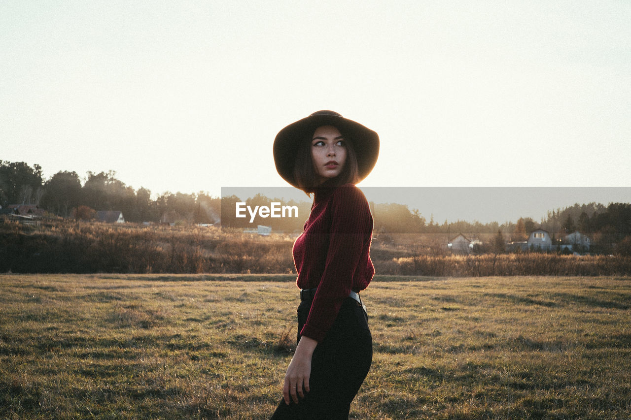 YOUNG WOMAN STANDING ON FIELD AGAINST SKY DURING SUNSET