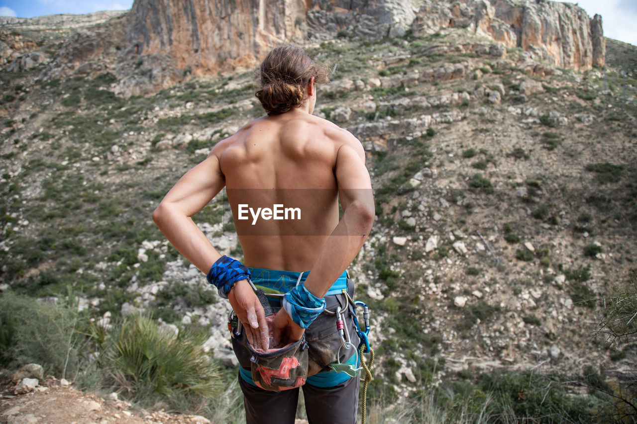 REAR VIEW OF SHIRTLESS MAN STANDING ON ROCK AGAINST MOUNTAIN