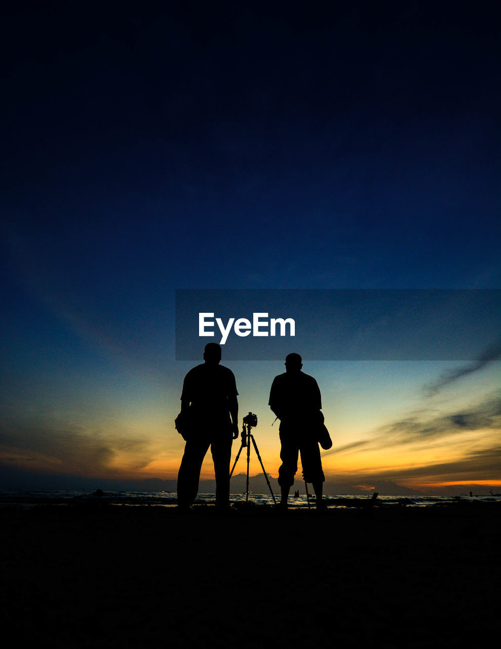 Full length of silhouette male friends standing amidst tripod at beach during sunset