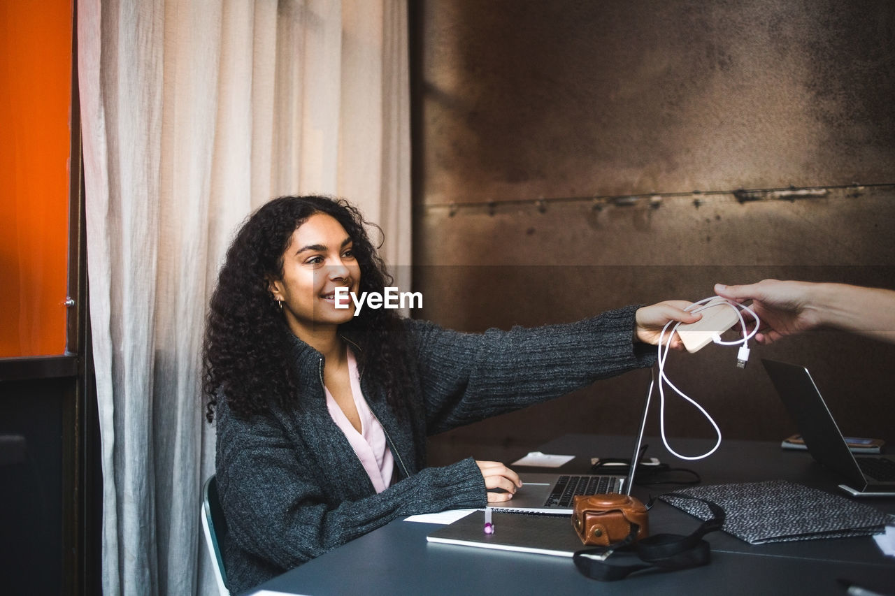 Smiling young female professional giving charger to male colleague at desk in office