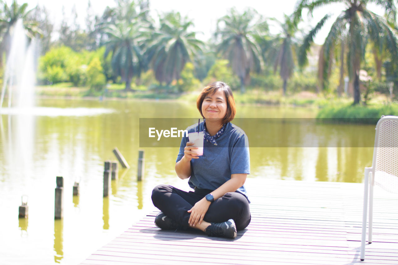 Full length of woman holding drink in glass while sitting on pier over lake against trees