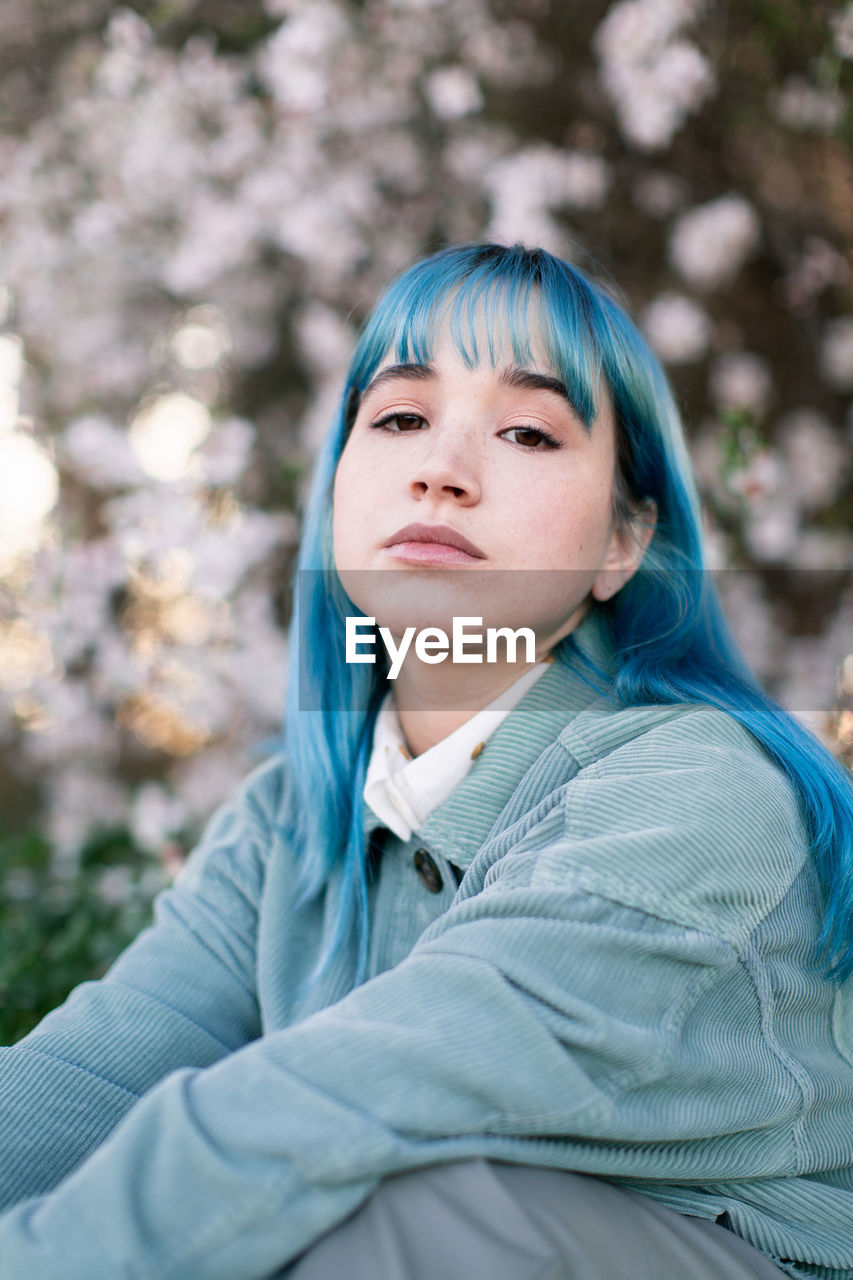 Sad millennial female model with blue hair in stylish outfit looking at camera thoughtfully while sitting on green grass near blooming tree in spring garden