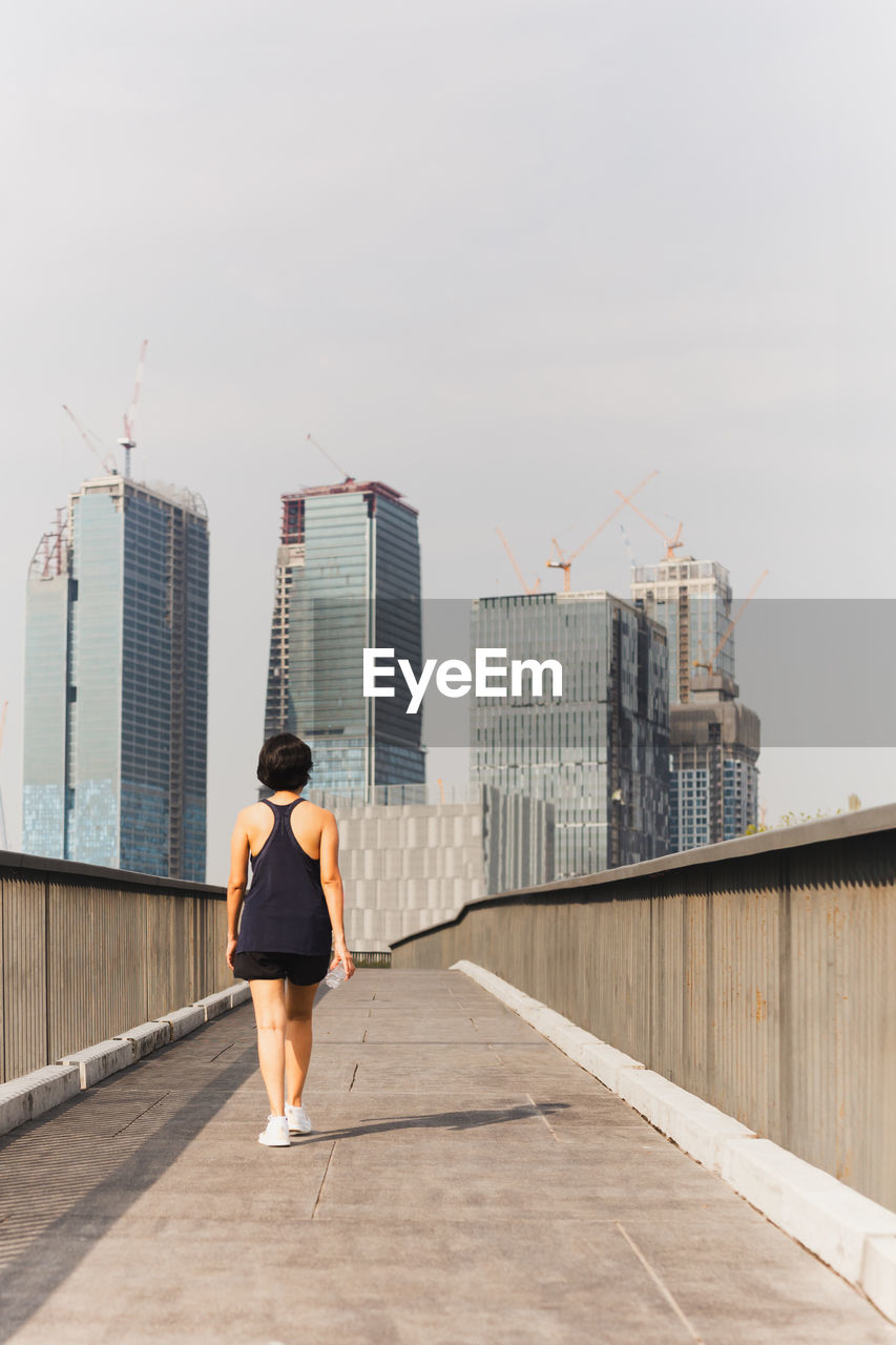 Woman exercise walking on the bridge hand holding bottle water with city building view.