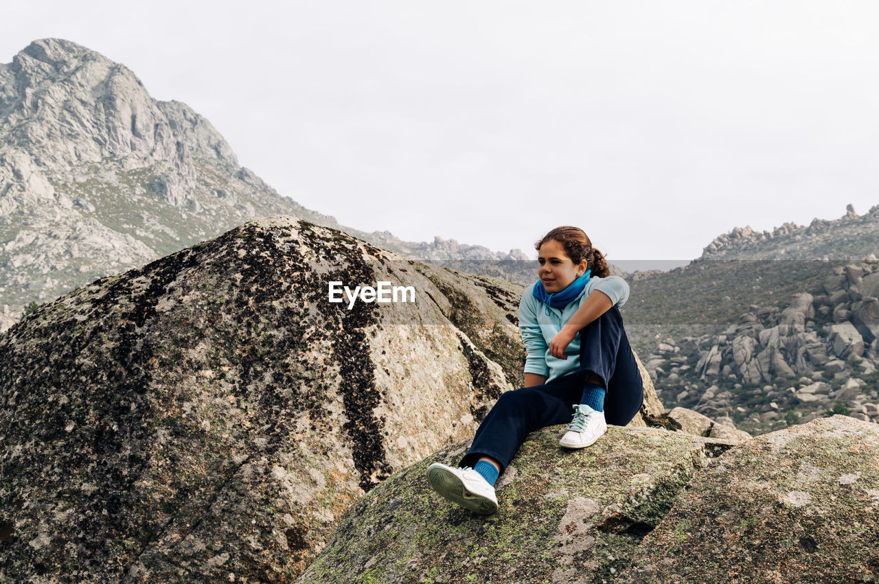 Thoughtful girl looking away while sitting on rock against clear sky