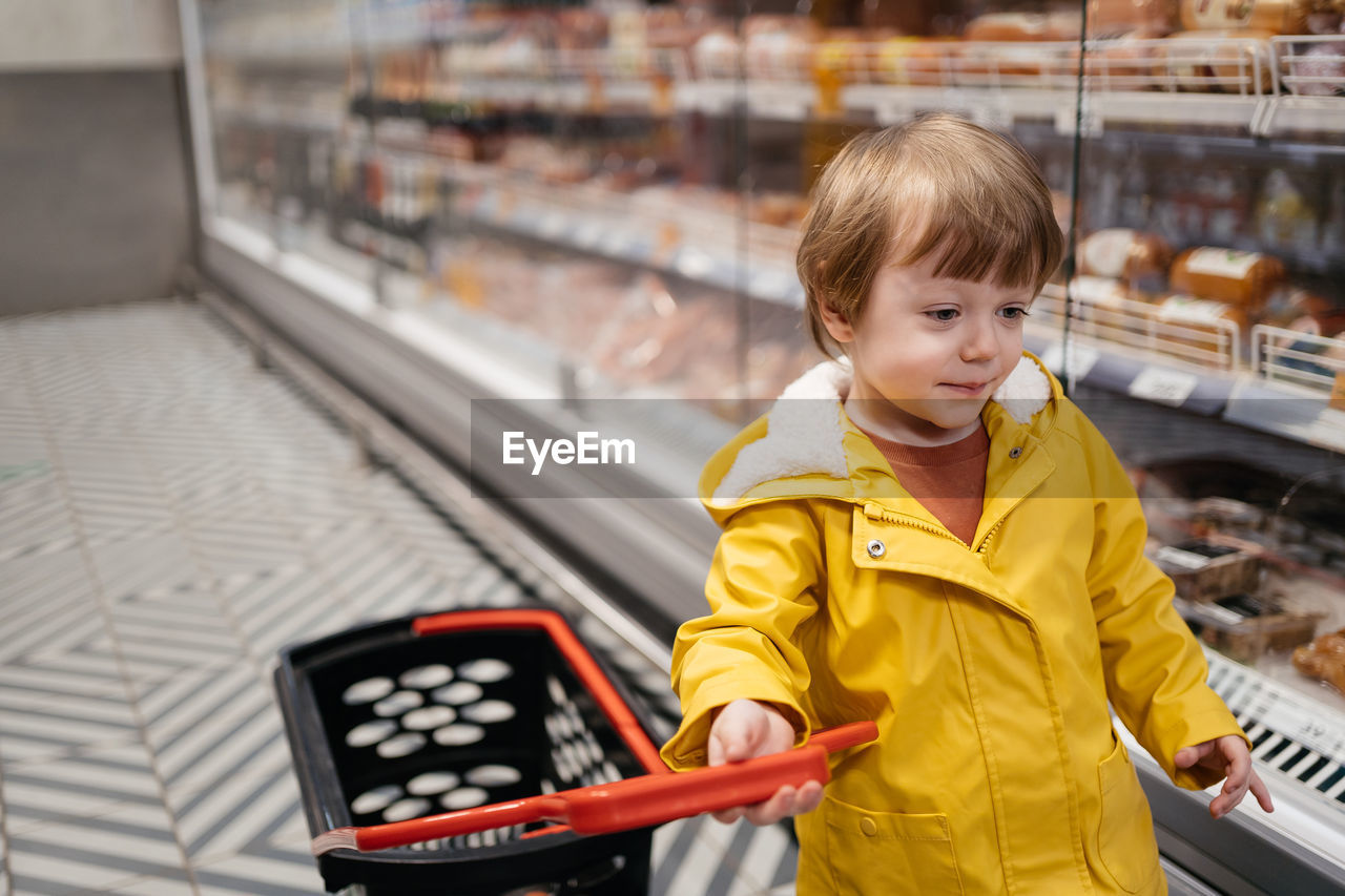 Child in the market with a grocery cart, wearing a yellow jacket and jeans