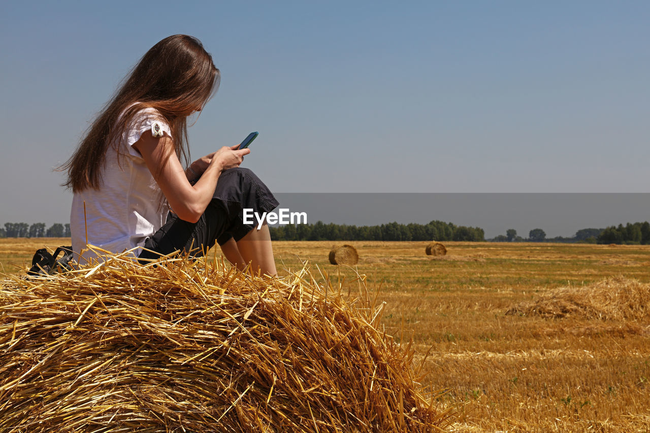 Side view of woman using mobile phone while sitting on hay bale at farm against clear sky
