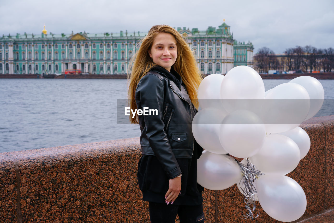 Caucasian girl holding white balloons standing by river on embankment of city