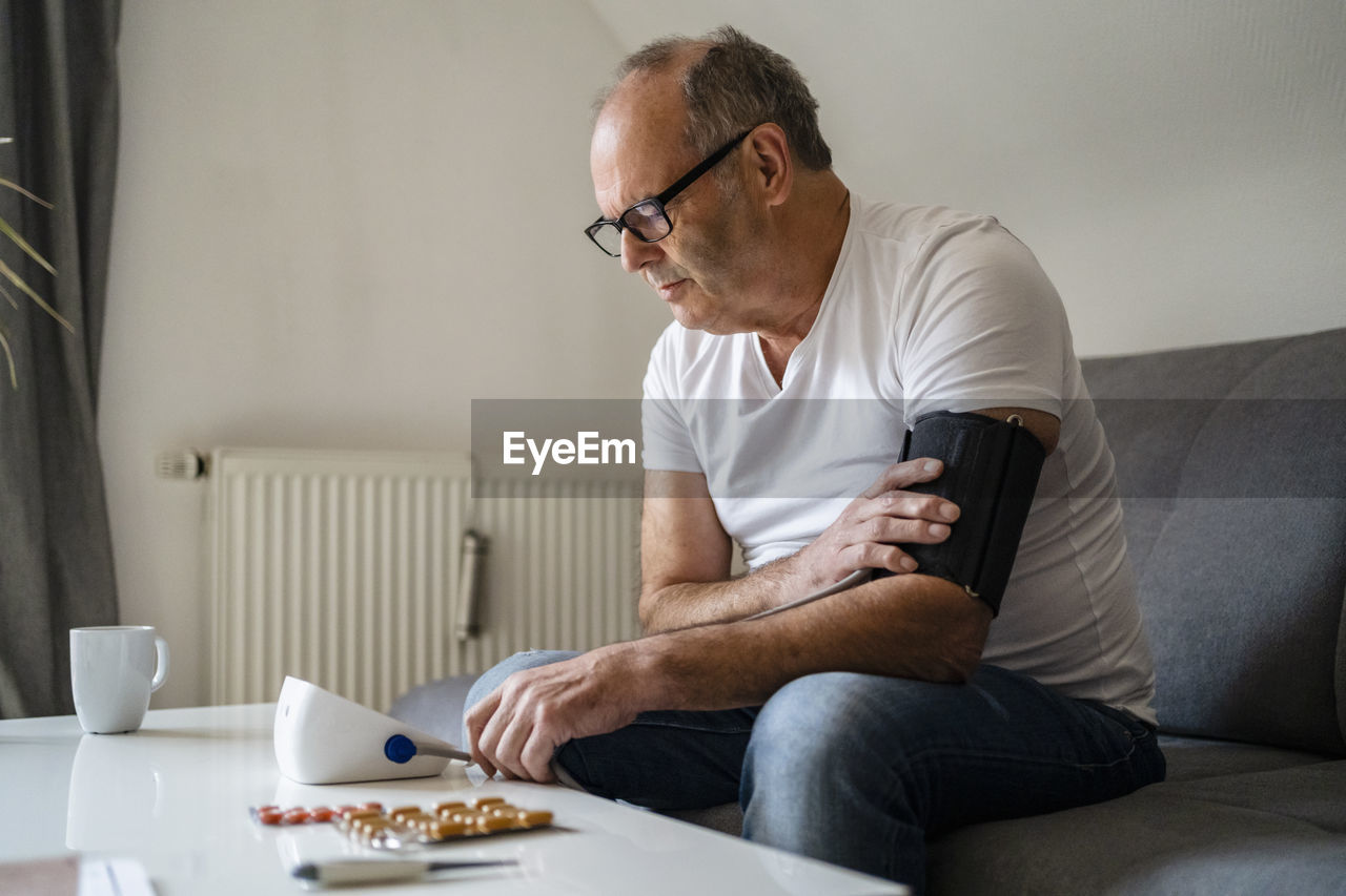 Senior man using blood pressure machine sitting on sofa at home